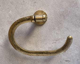 May include: A brass-colored, curved towel ring with an etched floral design. The ring is mounted on a circular base and has a rounded end. The metal has a polished finish, and the design is symmetrical.