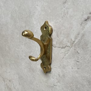 Op de afbeelding: Een messing wandhaak met een sierlijk ontwerp. De haak heeft een gebogen arm en een afgeronde knop aan het uiteinde. De montageplaat is rechthoekig met decoratieve uitsparingen. De haak is gemaakt van messing en heeft een gepolijste afwerking.