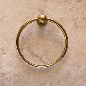 May include: A gold-colored towel ring with a circular design. The ring is mounted on a circular base with a small ball on top. The background is a light beige marble-like surface.