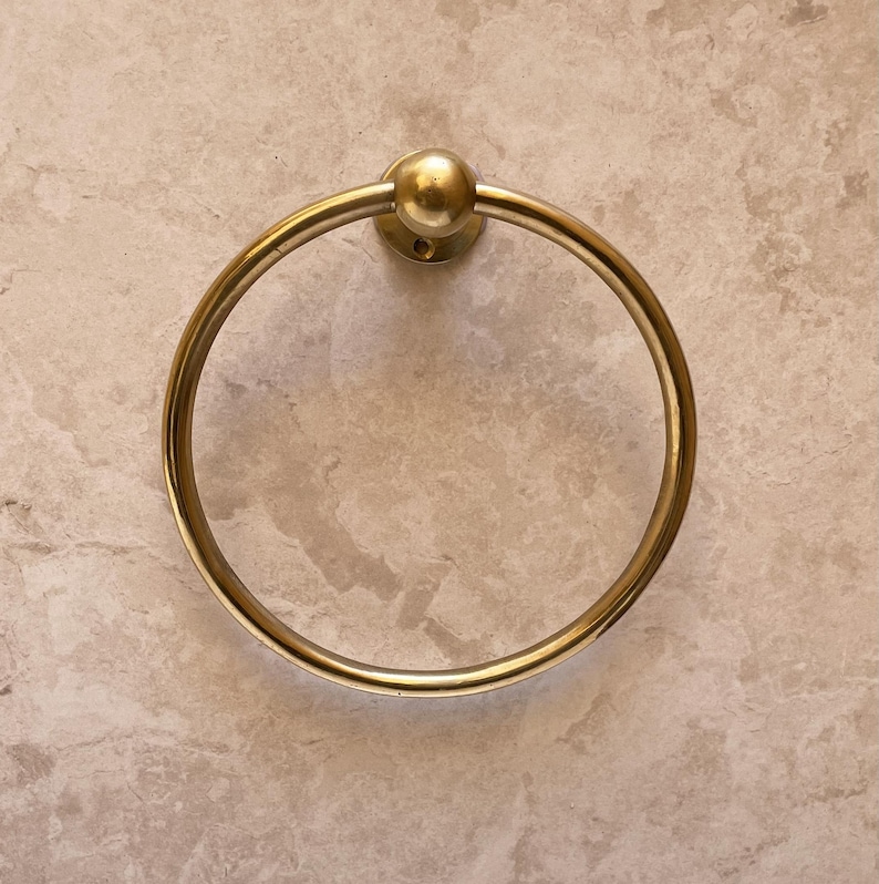 May include: A gold-colored towel ring with a circular design. The ring is mounted on a circular base with a small ball on top. The background is a light beige marble-like surface.