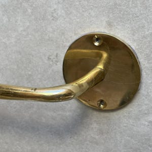 May include: A brass towel bar with a circular wall mount. The bar curves outward and is attached to the wall mount with a curved arm. The wall mount has two screw holes. The finish is a warm, aged brass.