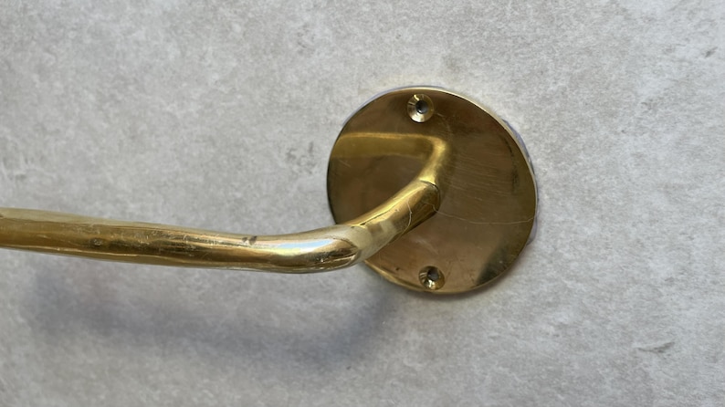 May include: A brass towel bar with a circular wall mount. The bar curves outward and is attached to the wall mount with a curved arm. The wall mount has two screw holes. The finish is a warm, aged brass.