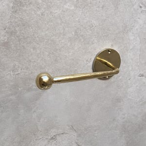 May include: A gold-colored, wall-mounted toilet paper holder. The holder features a circular backplate, a curved arm, and a spherical end. The fixture is made of metal and has a polished finish. The design is simple and modern.
