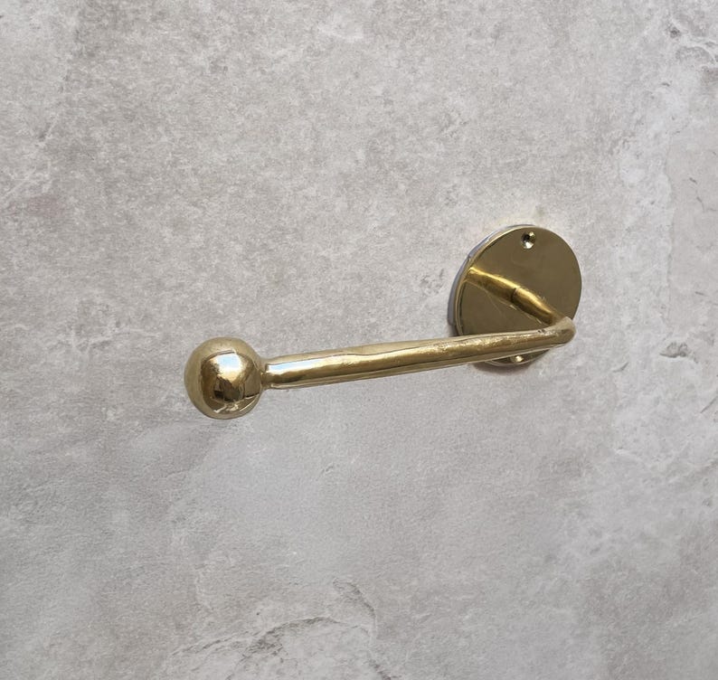 May include: A gold-colored, wall-mounted toilet paper holder. The holder features a circular backplate, a curved arm, and a spherical end. The fixture is made of metal and has a polished finish. The design is simple and modern.