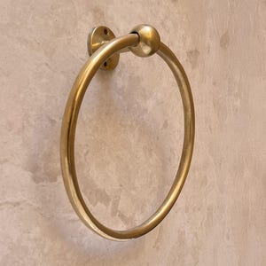 May include: A gold-colored towel ring with a circular design. The ring is mounted on a wall with a circular base and a ball-shaped detail at the top. The finish is shiny and reflective.