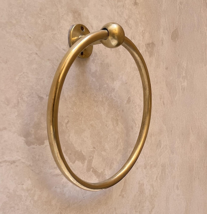 May include: A gold-colored towel ring with a circular design. The ring is mounted on a wall with a circular base and a ball-shaped detail at the top. The finish is shiny and reflective.