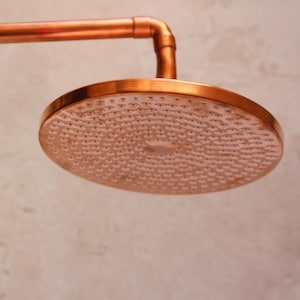 May include: A copper-colored shower head with a round, perforated spray surface. The shower head is attached to a copper pipe and elbow joint. The design is industrial and modern, with a focus on the warm tones of the metal.