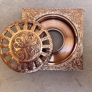 Pode incluir: Uma caixa decorativa de cor cobre com uma tampa aberta. A tampa é redonda com um intrincado design de explosão solar. A base quadrada e a tampa apresentam padrões em relevo detalhados. A caixa é provavelmente usada para guardar pequenos objetos.