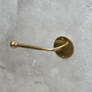 May include: A brass towel bar with a round mounting plate and a curved arm. The bar is mounted on a light gray stone wall. The bar is approximately 7 inches long.