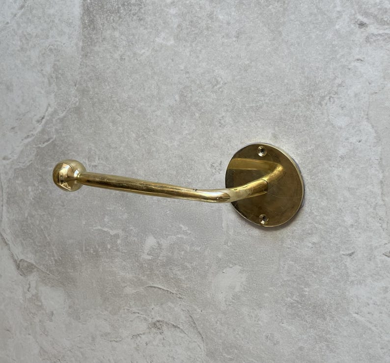 May include: A brass towel bar with a round mounting plate and a curved arm. The bar is mounted on a light gray stone wall. The bar is approximately 7 inches long.