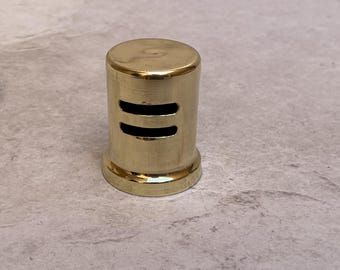 May include: A cylindrical, gold-colored bottle stopper with two horizontal, rectangular cutouts. The stopper has a rounded top and a flared base. The object is made of metal and has a shiny finish.