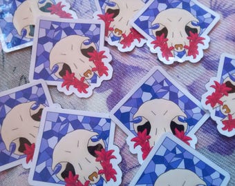 May include: A collection of square stickers featuring a white animal skull centred against a blue stained glass pattern, accented with bright red lily flowers. Each sticker displays the same graphic design with a white border and rounded corners.