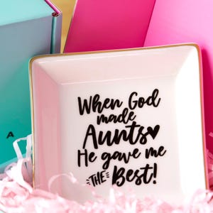 May include: A square, white ceramic trinket dish with a gold rim. The dish has the text "When God made Aunts He gave me THE Best!" in black script. The dish is surrounded by pink tissue paper.