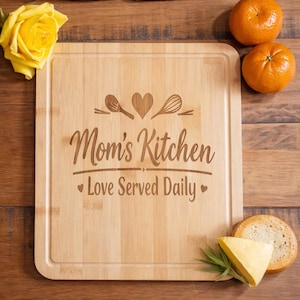 Op de afbeelding: Een houten snijplank met de tekst "Mom's Kitchen Love Served Daily" en keukengerei. Een mes, twee sinaasappels, een gele roos, kaas en brood zijn rond de plank gerangschikt.