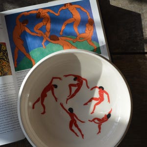 Könnte beinhalten: Weiße Keramikschale mit roter Illustration tanzender Figuren. Die Schale steht auf einer Holzoberfläche neben einem offenen Buch mit einem farbenfrohen Gemälde nackter Figuren. Das Bild hat warmes, natürliches Licht.