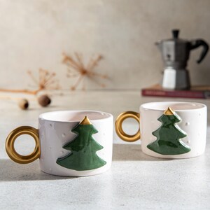 May include: Two white ceramic mugs with gold handles and a green Christmas tree design. The tree has a gold top. The mugs have small white dots and are on a light-colored surface. A silver coffee maker is in the background.