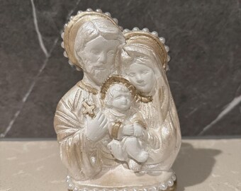 May include: A religious figurine depicting the Holy Family: Joseph, Mary, and baby Jesus. The statue is primarily white with gold accents, including halos and trim. The base is also gold, and the figurine is adorned with small, white pearl-like beads.