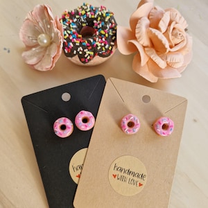 Puede incluir: Joyas y decoración con temática de donuts. Dos pares de pendientes de botón con forma de donut rosa sobre tarjetas negras y beige. Un donut glaseado con chocolate y chispas y dos flores de papel color melocotón también están en la imagen.