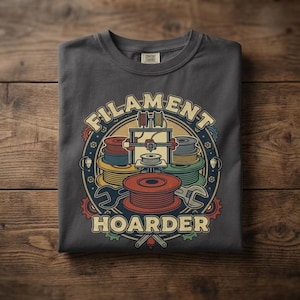 May include: Dark gray t-shirt with a circular graphic featuring a 3D printer, filament spools, and the words "FILAMENT HOARDER." The design includes wrenches and gears in a colorful, cartoon style. The shirt is folded on a wooden surface.