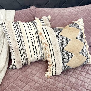May include: Two decorative throw pillows with geometric patterns. One pillow features black triangles and lines on a cream background, with fringe and pom-pom accents. The other has a black and cream zigzag design with fringe.