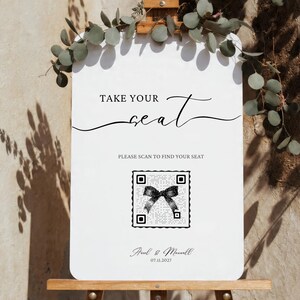 Puede incluir: Un letrero de boda blanco con el texto "TAKE YOUR seat" en escritura elegante. Debajo, dice "PLEASE SCAN TO FIND YOUR SEAT" con un c&oacute;digo QR. El letrero est&aacute; decorado con vegetaci&oacute;n y los nombres "Paul & Maxwell" y la fecha 07.11.2027.