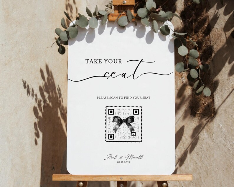 Puede incluir: Un letrero de boda blanco con el texto "TAKE YOUR seat" en escritura elegante. Debajo, dice "PLEASE SCAN TO FIND YOUR SEAT" con un c&oacute;digo QR. El letrero est&aacute; decorado con vegetaci&oacute;n y los nombres "Paul & Maxwell" y la fecha 07.11.2027.