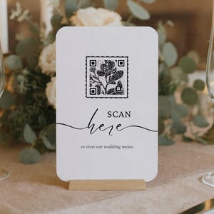 Puede incluir: Una tarjeta de men&uacute; de boda blanca con un c&oacute;digo QR y un dise&ntilde;o floral, sostenida en un soporte de madera. La tarjeta dice "ESCANEAR aqu&iacute; para ver nuestro men&uacute; de boda" en una escritura elegante. El fondo incluye flores y velas.