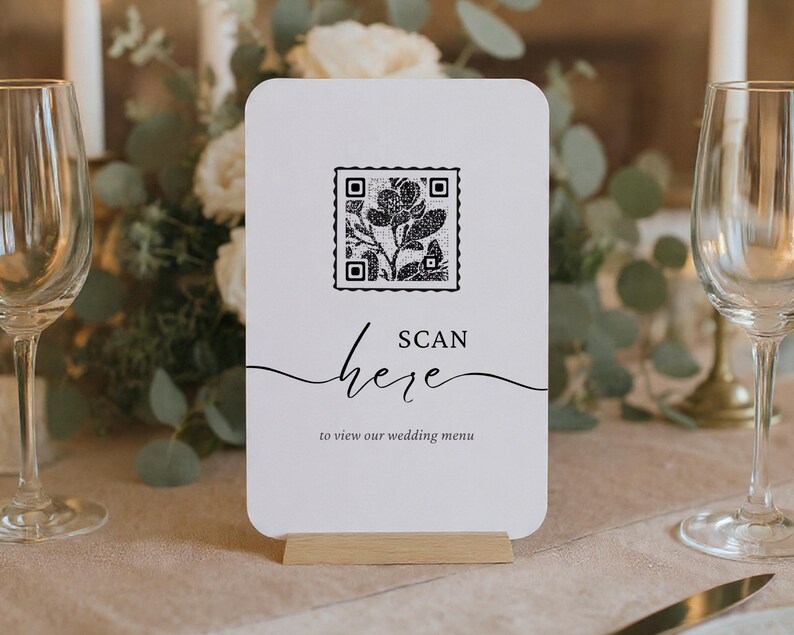 Puede incluir: Una tarjeta de men&uacute; de boda blanca con un c&oacute;digo QR y un dise&ntilde;o floral, sostenida en un soporte de madera. La tarjeta dice "ESCANEAR aqu&iacute; para ver nuestro men&uacute; de boda" en una escritura elegante. El fondo incluye flores y velas.