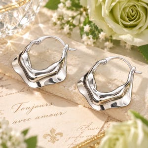 May include: A pair of silver-toned hoop earrings featuring a distinct, irregular, wavy, and rippled edge design. The earrings have a polished, reflective metallic finish and a hinged closure mechanism, showcasing a fluid, organic shape.