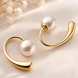 May include: A pair of gold-toned earrings featuring a curved, open-ended metal design. Each earring is accented with a single, smooth, round white pearl positioned at one end of the polished, sweeping gold-colored metal arc.