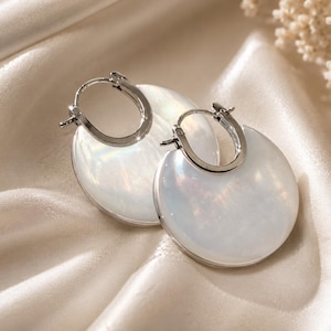 May include: A pair of hoop earrings featuring a polished silver-tone metal frame and a large, circular, iridescent white inlay. The earrings have a hinged closure mechanism at the top, showcasing a smooth, reflective surface with subtle pearlescent tones.
