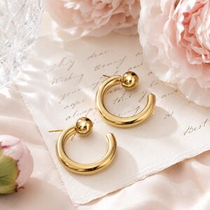 May include: A pair of gold-toned hoop earrings featuring a smooth, circular band design with a polished spherical bead accent at the top of each hoop. The earrings are constructed with a post-back closure mechanism for secure attachment.