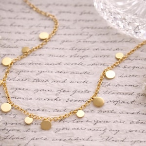 Può includere: Una delicata collana d'oro con ciondoli circolari. La collana è esposta su uno sfondo in stile vintage con scritte, circondata da fiori bianchi e una rosa rosa. Una ciotola di vetro trasparente è visibile.