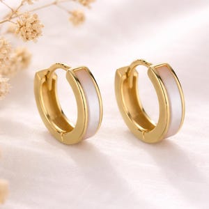 May include: A pair of gold hoop earrings featuring a smooth, white inlay along the outer edge. These small, circular earrings have a hinged closure mechanism and a polished metallic finish, presenting a clean and simple design for everyday wear.
