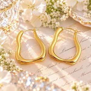 May include: A pair of gold hoop earrings with an irregular, wavy, organic shape. The earrings feature a polished, reflective surface and a hinged post closure. The design is asymmetrical, with smooth, flowing curves that create a unique silhouette.