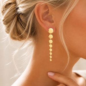 May include: A gold-toned drop earring featuring a vertical chain of eight circular discs that decrease in size from the top to the bottom. The earring has a polished, reflective surface and is attached to the earlobe with a circular stud component.