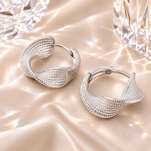 May include: A pair of silver-toned hoop earrings featuring a twisted, textured design with a fine cross-hatched pattern. The earrings have a hinged closure mechanism and a polished metallic finish that reflects light across the curved surfaces.