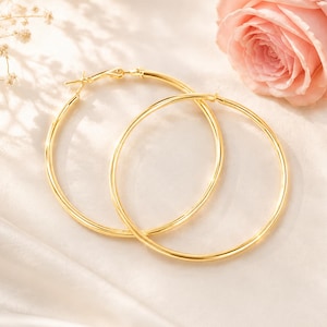 May include: A pair of large, polished gold hoop earrings with a smooth, circular design. The earrings feature a classic hinged post closure mechanism at the top, showcasing a bright, reflective metallic finish that catches the light.