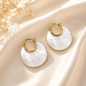 May include: A pair of gold hoop earrings featuring circular white mother-of-pearl pendants. The earrings have a polished gold metal finish and a smooth, iridescent white disc design that hangs from a small, rounded gold hoop closure.