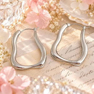 May include: A pair of silver-toned hoop earrings with an irregular, wavy, organic shape. The earrings feature a polished, reflective metallic finish and a hinged post closure mechanism, showcasing a fluid, non-symmetrical design.