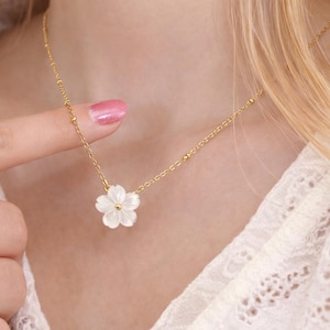 Puede incluir: Un delicado collar dorado con un colgante de flor blanca. La cadena es fina, con pequeñas cuentas espaciadas. El colgante floral tiene cinco pétalos y un centro redondo. El collar se lleva alrededor del cuello.