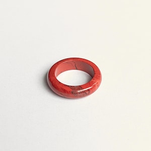 Könnte beinhalten: Ein roter Ring mit marmoriertem Aussehen. Der Ring ist ein massives Band mit glatter, polierter Oberfläche. Die Farbe ist ein leuchtendes Rot mit dunkleren, gesprenkelten Mustern. Ein schlichtes, aber elegantes Schmuckstück.