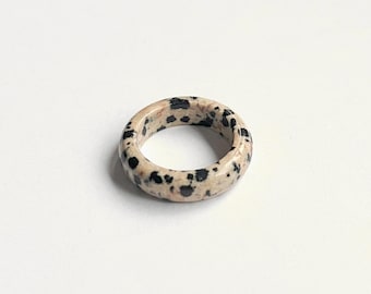 May include: A round ring made of speckled beige stone. The ring features a pattern of black spots across its surface. The ring is set against a plain white background, highlighting its natural colors and design.