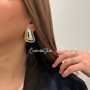 May include: Silver and gold-tone earrings with a unique triangular shape. The earrings have a gold-tone border with a textured design. The center of the earrings is silver. The image also shows multiple rings on a hand.