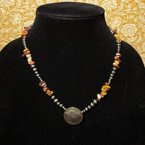 May include: A beaded necklace with a silver pendant featuring a buffalo. The necklace has small, round silver beads and colorful, irregularly shaped stone beads in shades of red, yellow, and white. The necklace is displayed on a black mannequin.