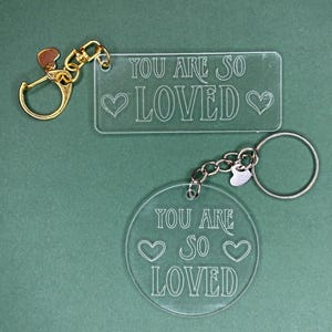 Peut inclure: Porte-clés en acrylique transparent avec les mots "YOU ARE SO LOVED" et des motifs de cœur. Un porte-clés est rectangulaire, l'autre est rond. Les deux ont du matériel argenté ou doré.