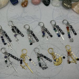 May include: A collection of ten metal keychains featuring silver or gold-toned lobster clasps, assorted gemstone beads, and charms including swords, daggers, bows, fans, and hearts. Some keychains include letter beads spelling BE YOU or LOVE.