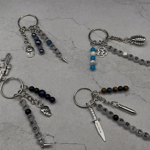 May include: Four metal keychains featuring character-themed charms and beads. One has a cap charm and beads spelling SGT GAZ. Another has a grenade charm and beads spelling SGT SOAP. A third has a rifle charm and beads spelling LT GHOST. The last has a knife charm and beads spelling CAP PRICE.