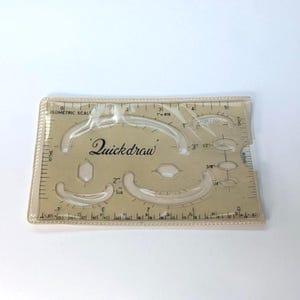Puede incluir: Una regla Quickdraw isométrica de plástico transparente con varias formas curvas y geométricas, y medidas en pulgadas. El texto "Quickdraw" está impreso en cursiva negra. La regla mide aproximadamente 15 cm de largo.