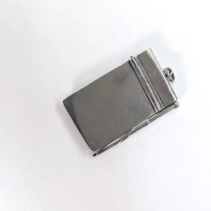 May include: A rectangular silver-toned metal charm shaped like a small notebook or case. The object features a smooth, flat surface with a hinged spine and a small metal loop at the top for attachment. A thin, pointed metal stylus is attached to the side.